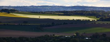 This landscape photograph, taken in Derbyshire, England, United Kingdom, features an agricultural scene with fields stretching across gently rolling hills. The main subject of the image is a sunlit section of farmland that stands out strikingly against the surrounding fields in deep shadow, creating a dramatic visual contrast. The photograph was captured in the early evening during the spring season, as indicated by the vibrant green of the crops and the lengthening shadows. The distant hills and layered countryside add depth to the view, emphasizing the rural character and agricultural productivity of this part of Derbyshire.